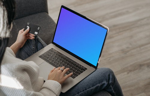 Mockup d'une utilisatrice sur un canapé faisant des achats en ligne avec son MacBook gris et une MasterCard à la main